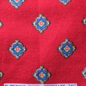 May include: Red fabric with a repeating pattern of ornate, diamond-shaped designs in blue, gold, and green. The fabric has a textured appearance. The text "MARCUS BROS. TEXTILES INC" is printed on a white strip.