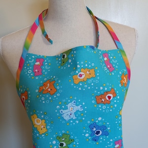 May include: A turquoise blue apron with a rainbow-colored strap. The apron features a repeating pattern of Care Bears on a blue background with white stars and hearts.