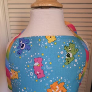 May include: A blue children's apron with a rainbow trim and a pattern of Care Bears on a blue background with white stars and hearts.