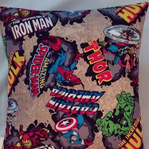 May include: Square pillow featuring comic book characters Iron Man, Spider-Man, Captain America, Thor, and Hulk. The pillow has a tan background with comic book panels and character names in bold, colorful text. The pillow is square.