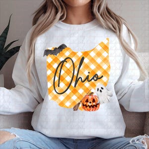 May include: White sweatshirt featuring a design with the state of Ohio in orange and yellow plaid. The word "Ohio" is written in black script. Halloween-themed graphics include a bat, pumpkin, and ghost.