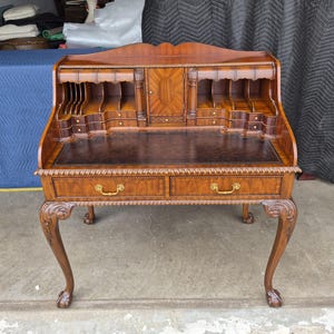 May include: A brown wooden secretary desk featuring a drop-front writing surface with a dark leather inlay, multiple interior cubby compartments, a central cabinet door, two lower drawers with gold-toned metal pulls, and four carved cabriole legs.