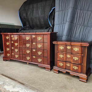 May include: A matching bedroom furniture set featuring a large dresser with eight drawers and a smaller nightstand with three drawers. Both pieces are made of dark wood and have ornate gold-colored hardware.