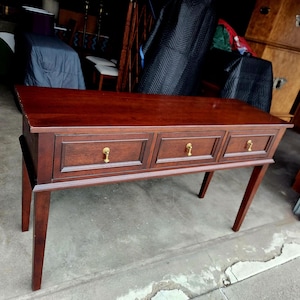 May include: A dark brown wooden console table with three drawers and gold-colored hardware. The table has four slender legs and a rectangular top. The drawers are evenly spaced across the front of the table.