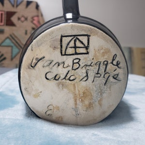 May include: Black ceramic mug with a white base. The base has a black square with a stylized letter A inside, and the text "Van Briggle Colo. Springs" in black cursive.