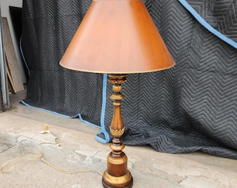 May include: A wooden table lamp with a brown lampshade and a decorative, carved wooden base. The base has gold accents and a round, ornate design. The lamp is plugged into an electrical outlet with a yellow cord.