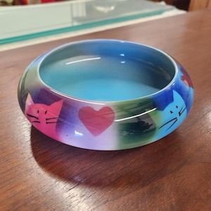 May include: A blue ceramic cat food bowl with a heart and two cat faces painted on it. The bowl has a blue, green, pink, and purple gradient.