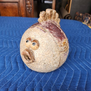 May include: A ceramic sculpture of a fish with a spiky fin, brown and white speckled body, and painted details. The fish has a large, round, brown eye and a small, open mouth.
