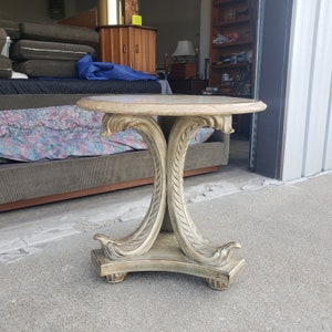 May include: A round, ornate side table with a light beige finish. The table has a carved, scrolled base with a decorative design. The table top is made of a light-colored material, possibly marble.