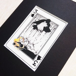 Pu&ograve; includere: Un'illustrazione in bianco e nero di una carta da gioco, il tre di picche. La carta presenta una donna con i capelli biondi sdraiata a terra, con una sagoma nera di un orso dietro di lei.