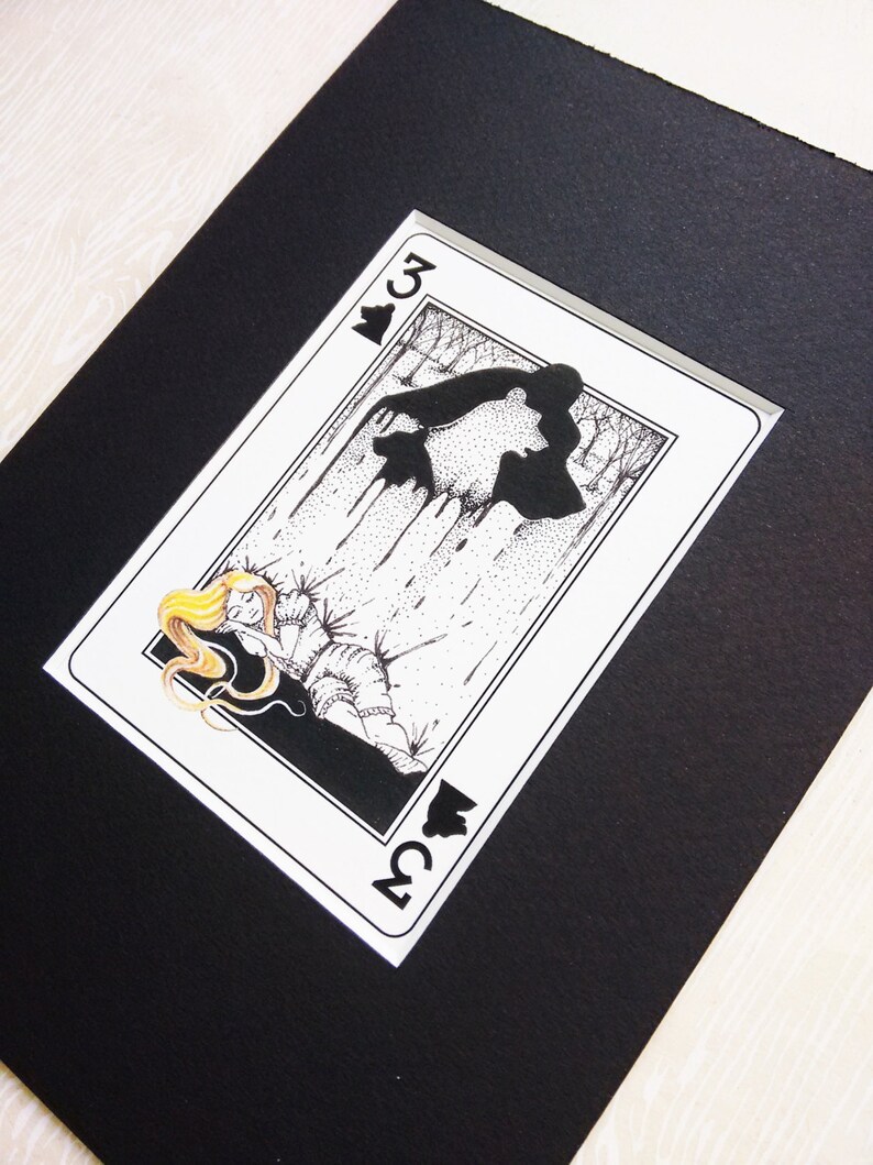 Pu&ograve; includere: Un'illustrazione in bianco e nero di una carta da gioco, il tre di picche. La carta presenta una donna con i capelli biondi sdraiata a terra, con una sagoma nera di un orso dietro di lei.