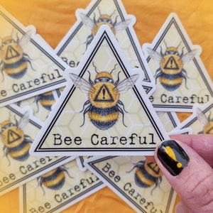May include: A set of yellow and black bee-themed stickers with the text "Bee Careful" on a white background. The stickers are on a yellow surface.