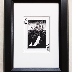 May include: A black and white illustration of a ship sailing on a starry sea with a whale jumping out of the water. The illustration is framed in a black frame and is part of a playing card deck. The card is the King of Spades.