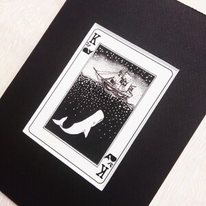 May include: A black and white illustration of a whale leaping out of the water with a ship sailing in the background. The illustration is on a playing card with the letter "K" and a spade symbol in the corner.