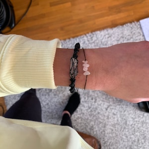 May include: Two bracelets on a wrist. One bracelet is black string with a clear crystal charm. The other bracelet is black string with three pink stone charms.