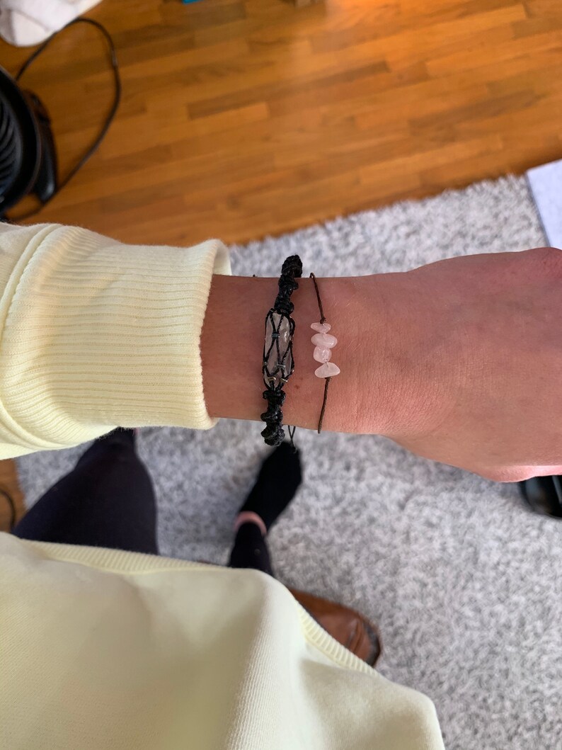 May include: Two bracelets on a wrist. One bracelet is black string with a clear crystal charm. The other bracelet is black string with three pink stone charms.
