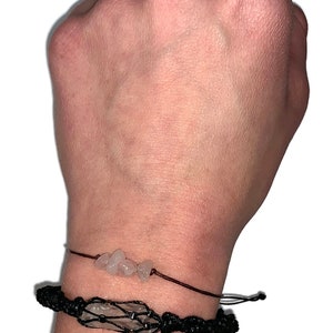 May include: Two bracelets on a wrist. The top bracelet is a simple string bracelet with small clear quartz stones. The bottom bracelet is a black macrame bracelet with a larger clear quartz stone in the center.