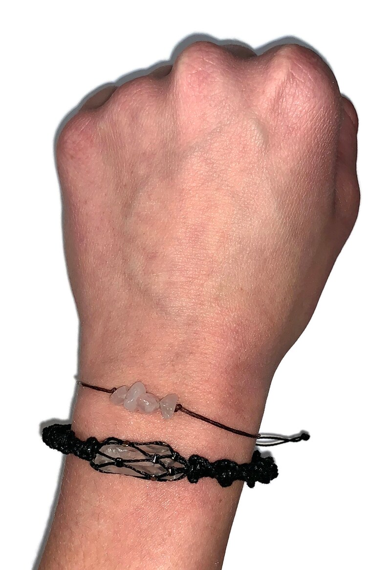 May include: Two bracelets on a wrist. The top bracelet is a simple string bracelet with small clear quartz stones. The bottom bracelet is a black macrame bracelet with a larger clear quartz stone in the center.