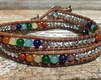 Op de afbeelding: Een meerstrengs lederen wikkelarmband met een zilveren sluiting. De armband heeft een verscheidenheid aan gekleurde edelsteen kralen, waaronder rood, oranje, geel, groen, blauw en paars.