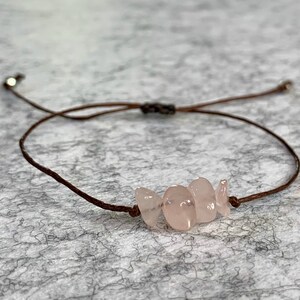 May include: A brown cord bracelet with a cluster of light pink rose quartz stones.