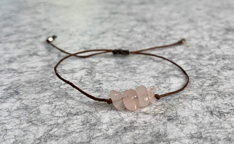 May include: A brown cord bracelet with a cluster of light pink rose quartz stones.