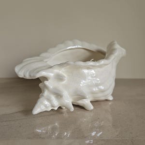 May include: White ceramic seashell planter with a smooth, glossy finish. The planter is shaped like a large seashell with a wide opening at the top.