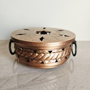 May include: A copper-coloured metal incense burner with a round shape and a lid. The lid has star-shaped and diamond-shaped cutouts. The body features a rope-like design and black handles.