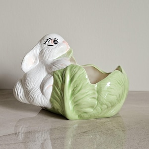 May include: A white ceramic bunny planter with pink detailing, set within a light green cabbage-shaped container. The bunny has detailed facial features and appears to be peeking out. A decorative Easter-themed item.
