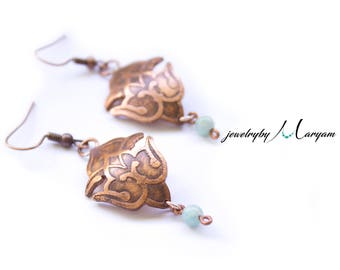 May include: A pair of copper-colored earrings with an ornate, floral design. Each earring features a small, light blue bead at the bottom. The earrings are displayed on a white background. The text "jewelry by Maryam" is visible.