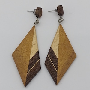 May include: A pair of wooden earrings with a geometric diamond shape. The earrings are made of three different types of wood: light brown, dark brown, and a light beige wood. The earrings have a silver hook.