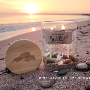 May include: A clear glass candle with the text "Lake Superior Beach Glass" and a wooden lid with a state outline. The candle contains decorative stones and sand. The image shows the candle on a sandy beach.