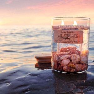 May include: A clear glass candle jar filled with Lake Superior agates and water, with two lit wicks. The label reads "Lake Superior Agates." The candle sits on a wet surface with a blurred sunset in the background.