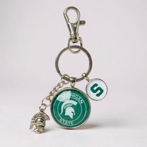 Puede incluir: Llavero plateado con múltiples colgantes. El colgante principal presenta las palabras "Michigan State" y un casco espartano en verde y blanco. Los colgantes adicionales incluyen un pequeño casco espartano y un colgante circular con la letra "S".