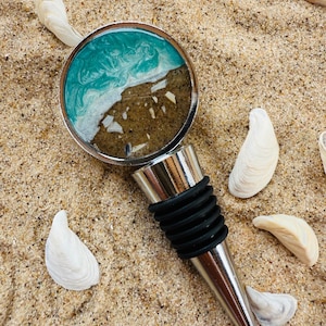 May include: A bottle stopper with a beach-themed design. The stopper has a silver-coloured metal base and a black rubber seal. The top features a teal and white wave design with sand and shell accents. White seashells are scattered around the stopper on a bed of sand.