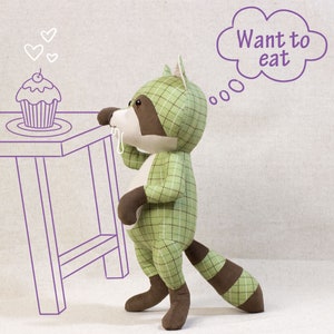 Puede incluir: Un juguete de peluche de mapache de cuadros verde y marr&oacute;n con una cola marr&oacute;n, mirando un cupcake con una cereza en la parte superior. El mapache tiene un globo de pensamiento sobre su cabeza que dice "Quiero comer".