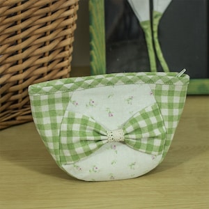 Pode incluir: Uma pequena bolsa de tecido vichy verde e branco com um laço branco e estampa floral. A bolsa tem um fecho de correr.