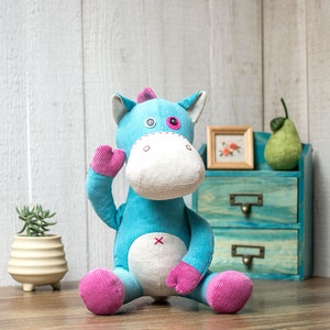 May include: A blue and pink plush toy shaped like a horse with a white face and a pink button nose. The toy is sitting on a wooden surface in front of a blue and white dresser.