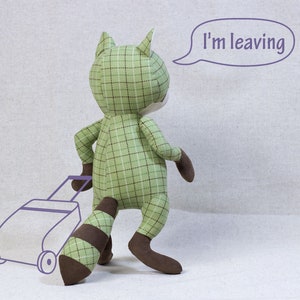 Puede incluir: Un juguete de peluche de mapache verde y marr&oacute;n con un patr&oacute;n de cuadros est&aacute; tirando de una peque&ntilde;a maleta con el texto "Me voy" en un globo de di&aacute;logo arriba.