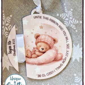 May include: A circular Christmas ornament featuring a sleeping teddy bear illustration in pink and white. The ornament has a ribbon and the text: "Untie this ribbon and you will see, just how small I used to be." From Unique & Crafty.