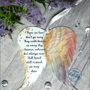 May include: White heart-shaped ornament with a painted angel wing design in shades of pink, yellow, and blue. The ornament has gold glitter accents and a white ribbon for hanging. The ornament features the text "Those we love don't go away. They walk beside us every day. Unseen, unheard but always near. Still loved, still missed. So very dear."