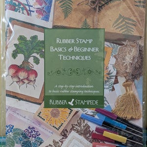 May include: A instructional book titled Rubber Stamp Basics & Beginner Techniques by Rubber Stampede, featuring a collage of stamped floral and vegetable designs, a gold tassel, and a set of colorful markers in a clear plastic protective sleeve.