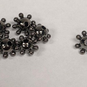 May include: A collection of dark gray metal bead caps, each with a central hole and small spheres radiating outwards. The bead caps are arranged on a white surface, with one cap isolated.