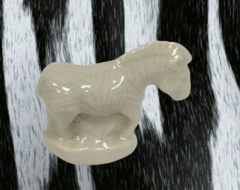 May include: A small, off-white ceramic horse figurine. The horse is in a standing position with its head slightly lowered. The figurine has a smooth, glossy finish and is set against a black and white zebra-striped background.