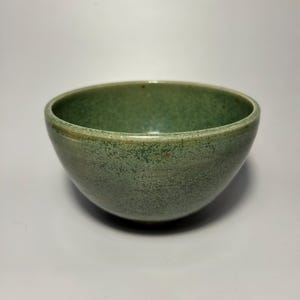 May include: A small, green ceramic bowl with a speckled glaze. The bowl is round and has a slightly flared rim.