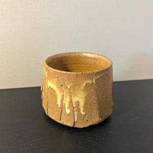 May include: A small, cylindrical ceramic cup with a speckled tan and brown glaze. The exterior features a textured surface with vertical indentations and a lighter, cream-colored drip pattern running down the sides from the rim.
