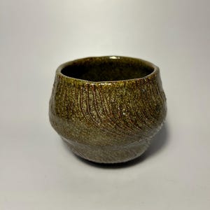 May include: A handmade ceramic cup with a textured surface. The cup has a wide, rounded base and a slightly flared rim. The exterior is a mottled olive green and brown color, with a glossy finish. The cup is suitable for beverages.