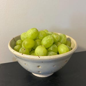 May include: A light gray ceramic bowl with a speckled finish and small circular indentations around the exterior, filled with a cluster of fresh, round, light green grapes. The bowl has a wide rim and a tapered base, holding the fruit pile.