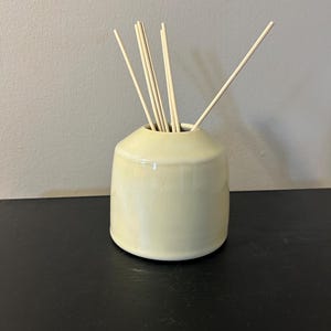 May include: A pale yellow ceramic reed diffuser vessel with a smooth, glossy finish and a tapered neck. Several thin, light-colored wooden reeds are inserted into the opening, extending upwards and outwards from the center of the container.