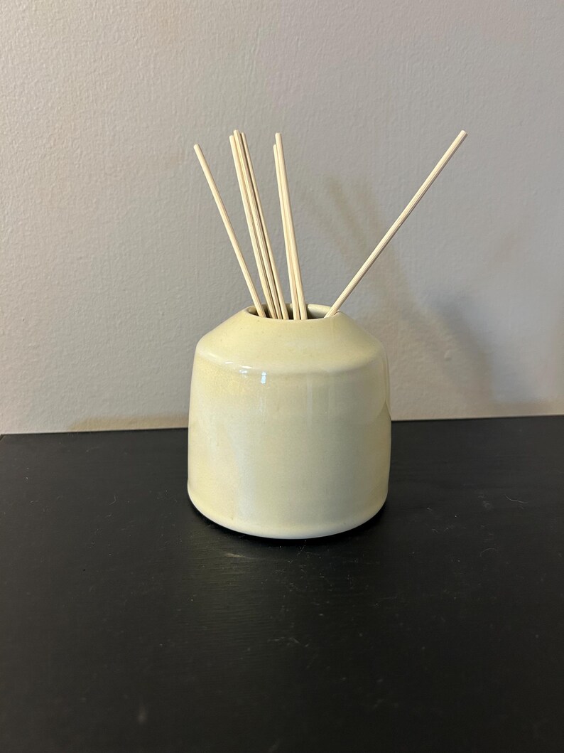 May include: A pale yellow ceramic reed diffuser vessel with a smooth, glossy finish and a tapered neck. Several thin, light-colored wooden reeds are inserted into the opening, extending upwards and outwards from the center of the container.