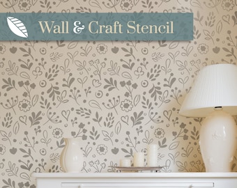Puede incluir: Una pared decorada con un patrón de plantilla floral en gris sobre un fondo beige. Las palabras "Wall & Craft Stencil" se muestran en una pancarta azul. Una cómoda, lámpara y velas blancas están en primer plano.